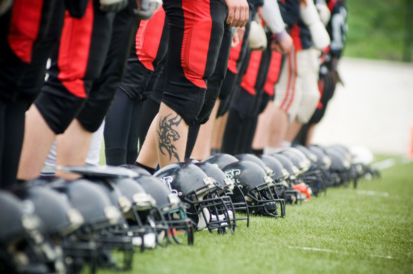 Photograph of a football team standing with their helmets on the grass in front of their feet - the photo shows the players from the waist down